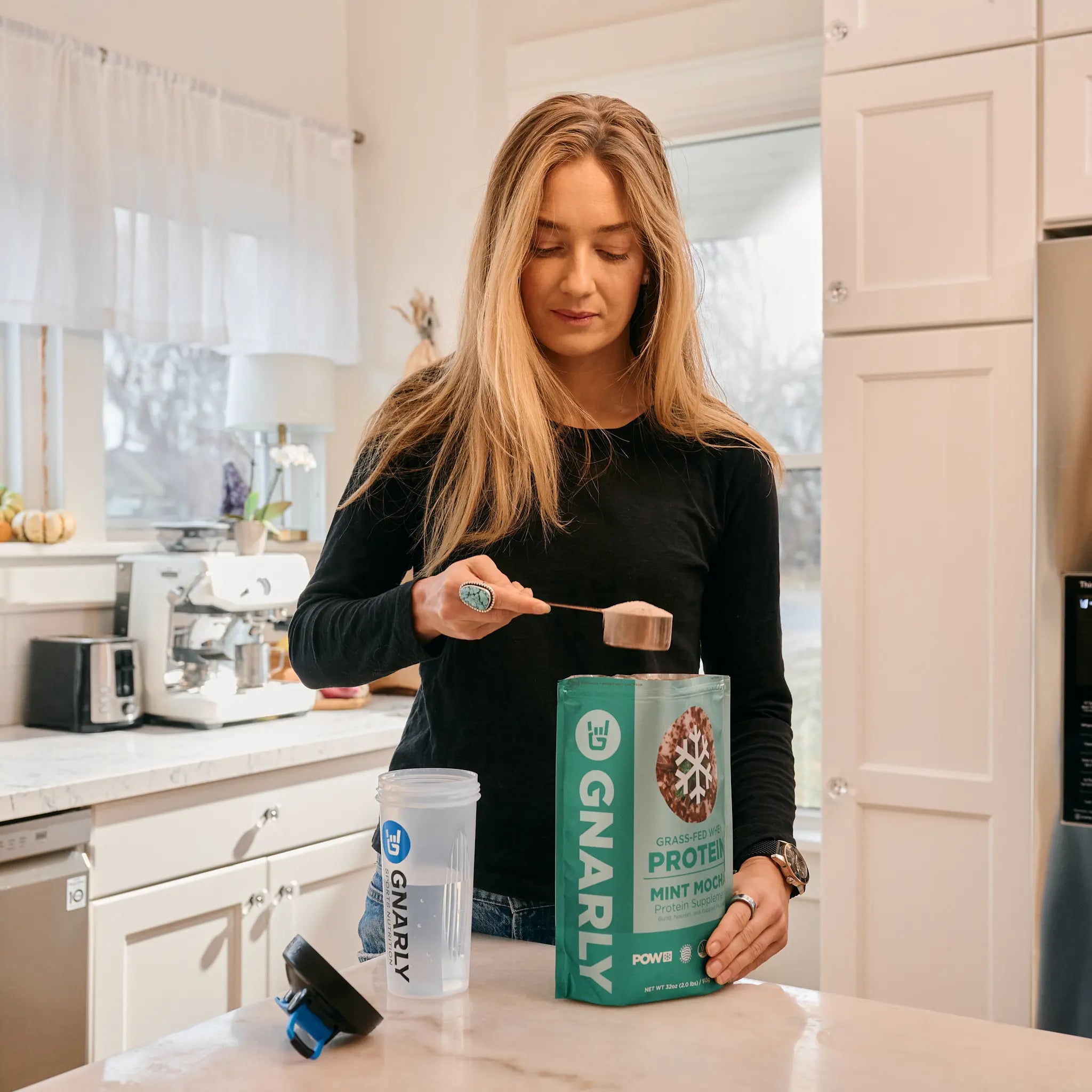 Woman in a kitchen holding GNARLY protein powder and a scoop.